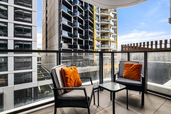 A balcony with two chairs, one with an orange cushion and another with a grey cushion, overlooking a modern building with multiple balconies. - Bright 2-bed Flat With Cityside Charm (Melbourne)