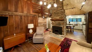 Un salon chaleureux avec une cheminée en pierre, des murs en bois, un fauteuil à carreaux, une table basse en bois ornée d’une plante, et une cuisine en arrière-plan.