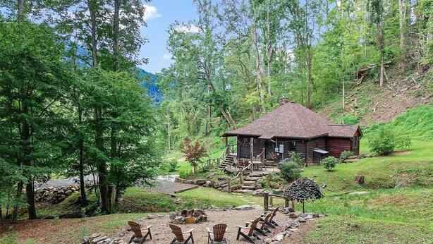 Un chalet rustique doté d’un foyer en pierre et de sièges d’extérieur, entouré d’une forêt dense et traversé par un petit ruisseau.