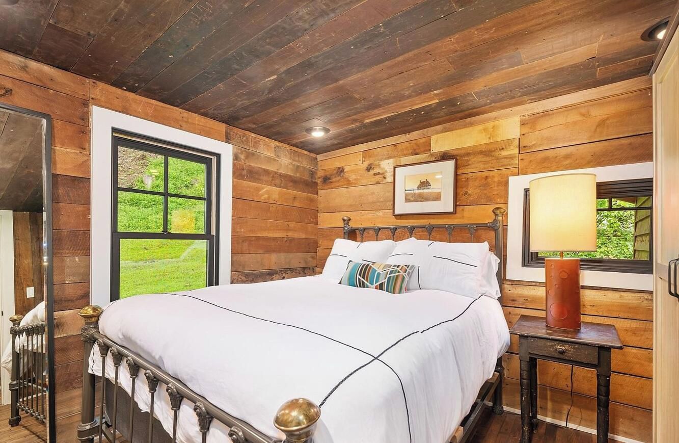 Un dormitorio con paneles de madera, una cama grande, una ventana con vistas a zonas verdes y una lámpara de noche.