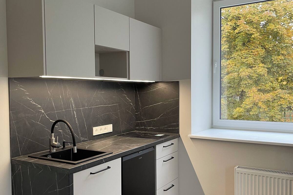 Een moderne keuken met zwarte marmeren aanrechtbladen, witte kasten en een raam met uitzicht op bomen.