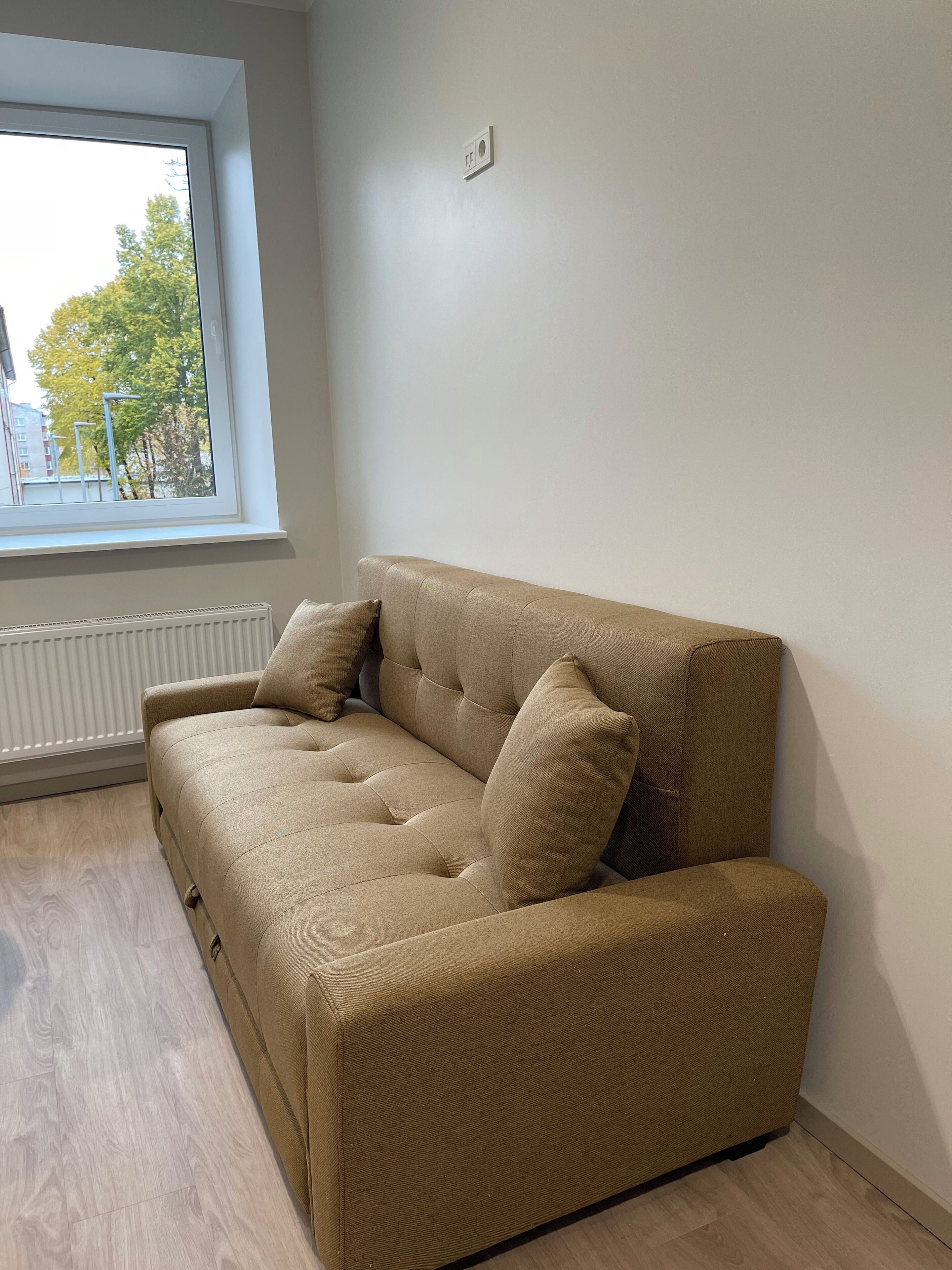 A beige sofa with two cushions in a room with a large window, white walls, and a radiator.