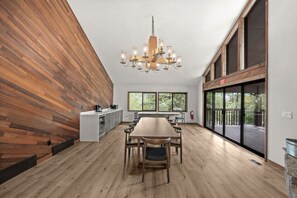 Une pièce spacieuse avec une longue table en bois, un lustre moderne et de grandes fenêtres donnant sur l’extérieur.