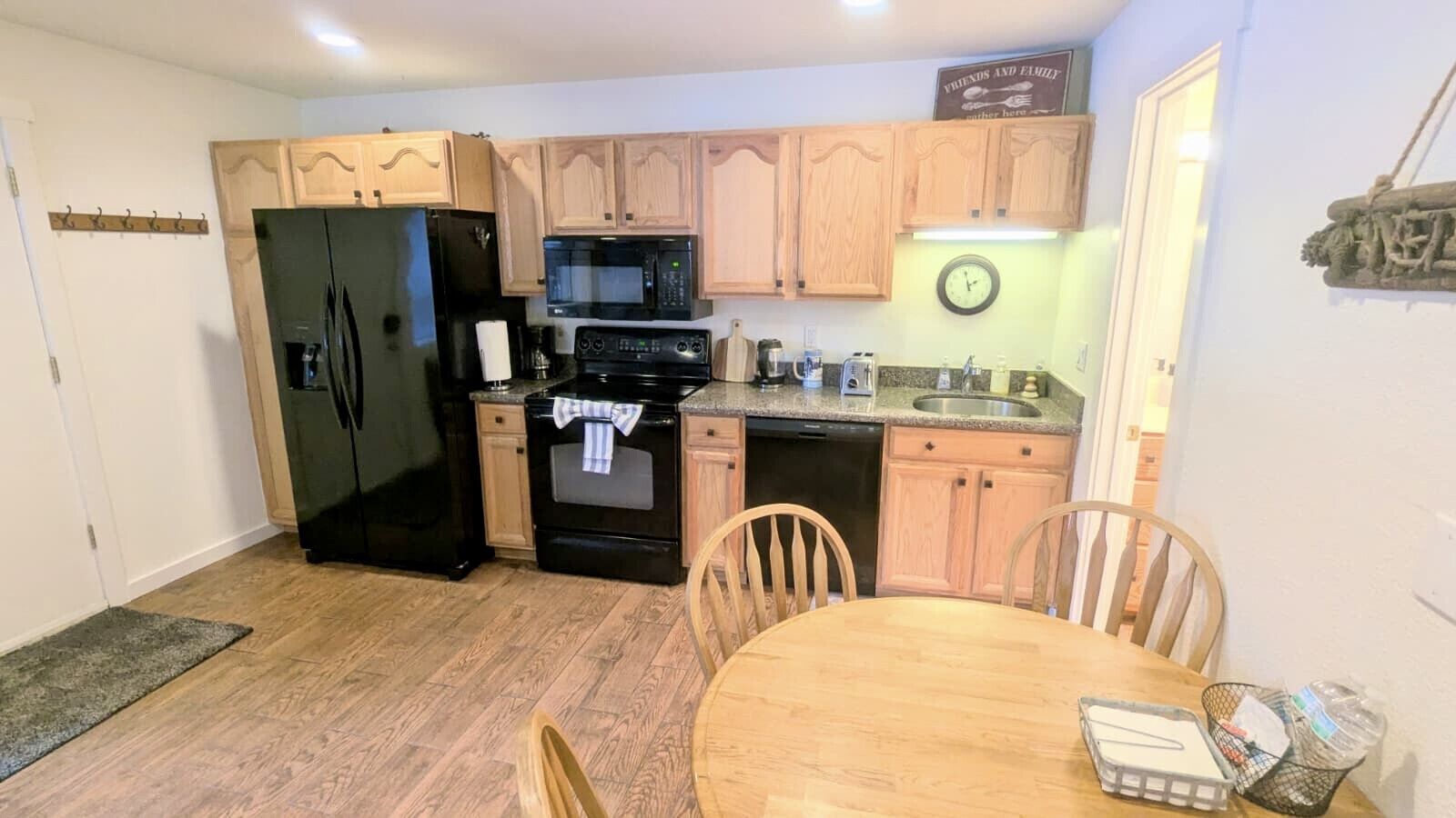 A compact kitchen with wooden cabinets, a black refrigerator, a microwave, an oven, and a sink. There is a dining table with chairs and a clock on the wall.