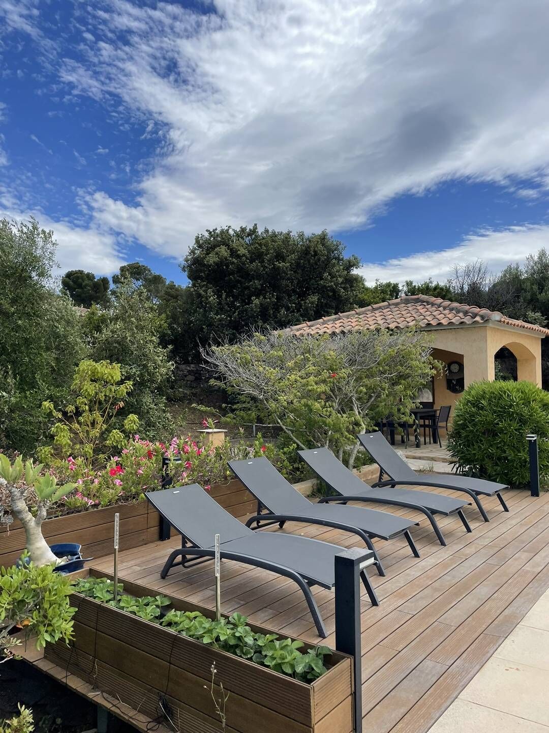 A patio with sun loungers, a wooden deck, and a garden with various plants and flowers.