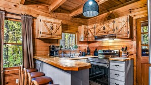 Une cuisine de style rustique, avec des meubles en bois, un îlot central, des tabourets de bar et une grande fenêtre donnant sur la forêt.
