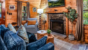 L’intérieur d’un chalet confortable, avec une cheminée, des murs en bois, un canapé agrémenté de coussins à motifs floraux et un téléviseur fixé au-dessus de la cheminée.