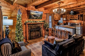 A cozy cabin interior with a fireplace, wooden walls, a dining area, and a living room with a sofa and chairs.