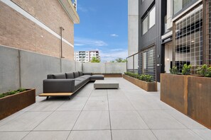 An outdoor seating area with a sofa and planters, a tiled floor, and a building with large windows and a brick wall.