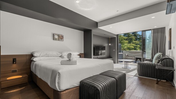 A modern hotel room with a large bed, a seating area with a sofa and armchairs, a television, and a view of the outdoors.