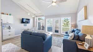 A modern living room with a sofa, a coffee table, a TV, and a ceiling fan. - Salty Blue Hen: Oceanside, Community Pool, Close to Beach Access (Corolla)
