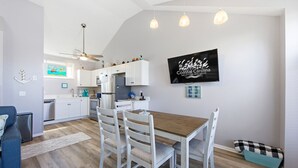 A modern kitchen with white cabinets, a wooden dining table, and a TV displaying 'Coastal Carolina Vacations and Sales'. - Salty Blue Hen: Oceanside, Community Pool, Close to Beach Access (Corolla)