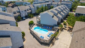 A residential area with a swimming pool surrounded by lounge chairs, a view of the ocean, and multiple apartment buildings. - Salty Blue Hen: Oceanside, Community Pool, Close to Beach Access (Corolla)