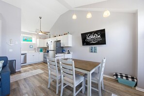 Una cocina moderna con armarios blancos, una mesa de comedor de madera y un televisor mostrando 'Coastal Carolina Vacations and Sales'.