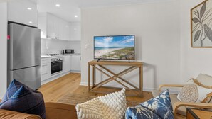 Una sala de estar moderna con una televisión de pantalla plana sobre un soporte de madera, un sofá con cojines estampados y una cocina al fondo.