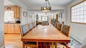 A spacious dining area with a long wooden table and chairs, a chandelier, and a view of the kitchen and living area. - Lakeshore Retreat- Estes Park Escape with Mountain & Pond Views (Estes Park)