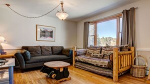 A cozy living room with a dark grey sofa, a patterned sofa, a wooden coffee table, and a large window with curtains. - Lakeshore Retreat- Estes Park Escape with Mountain & Pond Views (Estes Park)