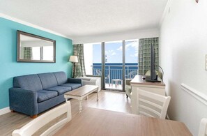 A room with a sofa, a table, a chair, a lamp, a TV, and a balcony with a view.