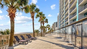 A wooden boardwalk with sun loungers, palm trees, and a building with balconies. - Caribbean 1020 (Myrtle Beach)