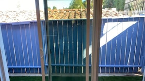 Una stanza con una porta scorrevole in vetro che si apre su un giardino sul retro, con recinzione blu e tetto in tegole.