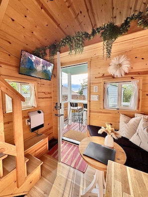 Een houten blokhutinterieur met een televisie, een bank, een eettafel en uitzicht op buiten.