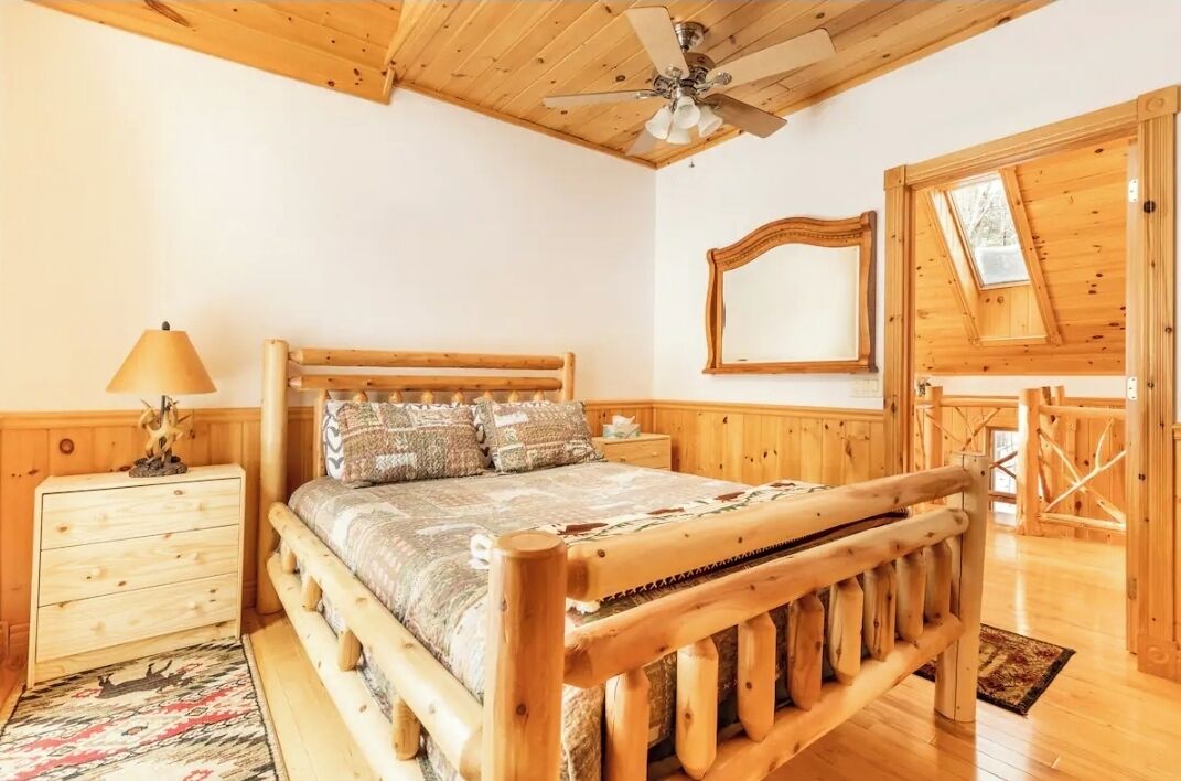 A wooden bedroom with a bed, a nightstand, a lamp, a ceiling fan, and a window with a view of the outside.