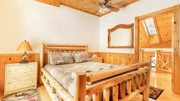 A wooden bedroom with a bed, a nightstand, a lamp, a ceiling fan, and a window with a view of the outside.