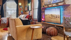 A cozy living room with a sofa, armchair, and ottoman, a flat-screen TV mounted on a brick wall, and a window with curtains. - Panther City Pad - Downtown Fort Worth (Fort Worth)