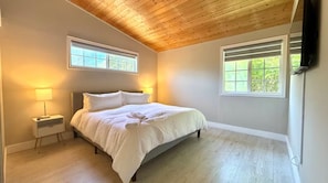 Un dormitorio con una cama, dos mesitas de noche, un buró, techo de madera y dos ventanas.