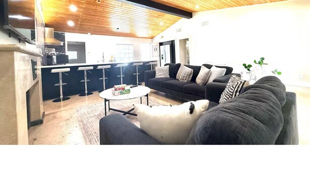 A modern living room with a wooden ceiling, a black sofa, a white coffee table, and a kitchen area in the background. - 3-bedroom house near Disneyland in Anaheim with AC (Anaheim)