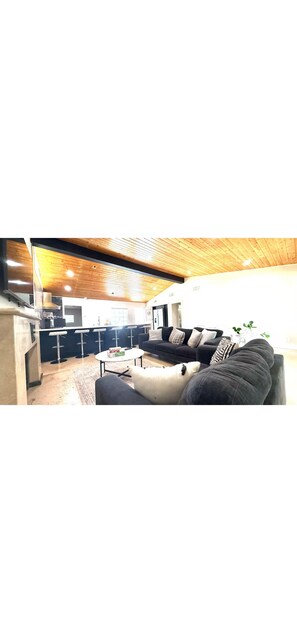 A modern living room with a wooden ceiling, a black sofa, a white coffee table, and a kitchen area in the background. - 3-bedroom house near Disneyland in Anaheim with AC (Anaheim)