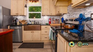 Cozinha com armários de madeira, lava-louças em aço inoxidável, janela com vista para vegetação e bancada preta com diversos utensílios de cozinha.