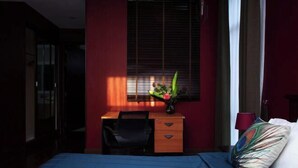 Un dormitorio con cama, escritorio y silla. Hay una ventana con persianas, un florero con flores y una lámpara en la mesita de noche.