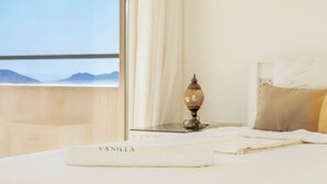A neatly made bed with a white blanket and a towel labeled "VANILLA" on a hotel room with a view of mountains through a large window.