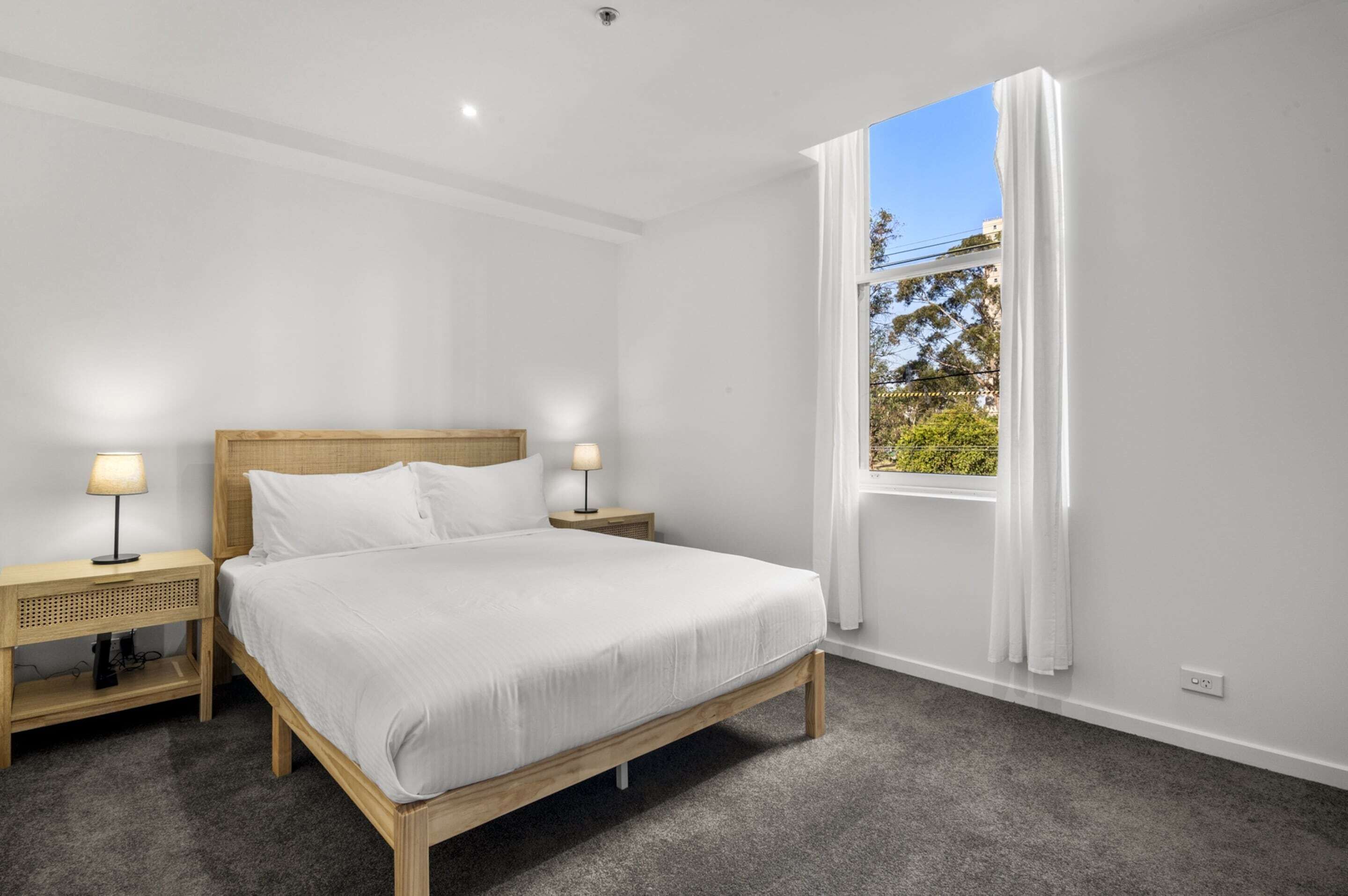 A bedroom with a large bed, two bedside tables with lamps, a window with curtains, and a view of trees outside.