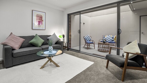 A modern living room with a grey sofa, a glass coffee table, and a sliding glass door leading to an outdoor seating area with a table and chairs.