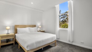 A bedroom with a wooden bed, two bedside tables with lamps, a large window with white curtains, and a view of greenery outside.
