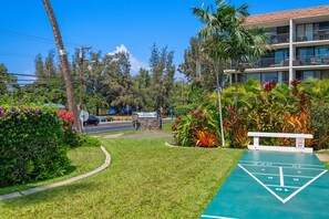 A well-maintained grassy area with a shuffleboard court, surrounded by lush tropical plants and a multi-story building with balconies.