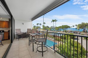 A balcony with a dining area, a pool, and a view of the ocean and palm trees.