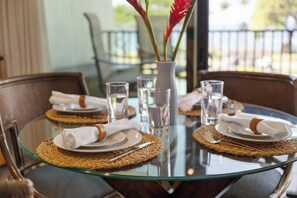 A dining table with a glass top, surrounded by chairs with woven placemats and neatly folded white napkins.