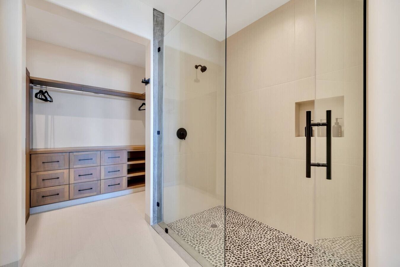 A modern bathroom with a glass shower enclosure, a walk-in closet with wooden drawers, and a tiled floor.