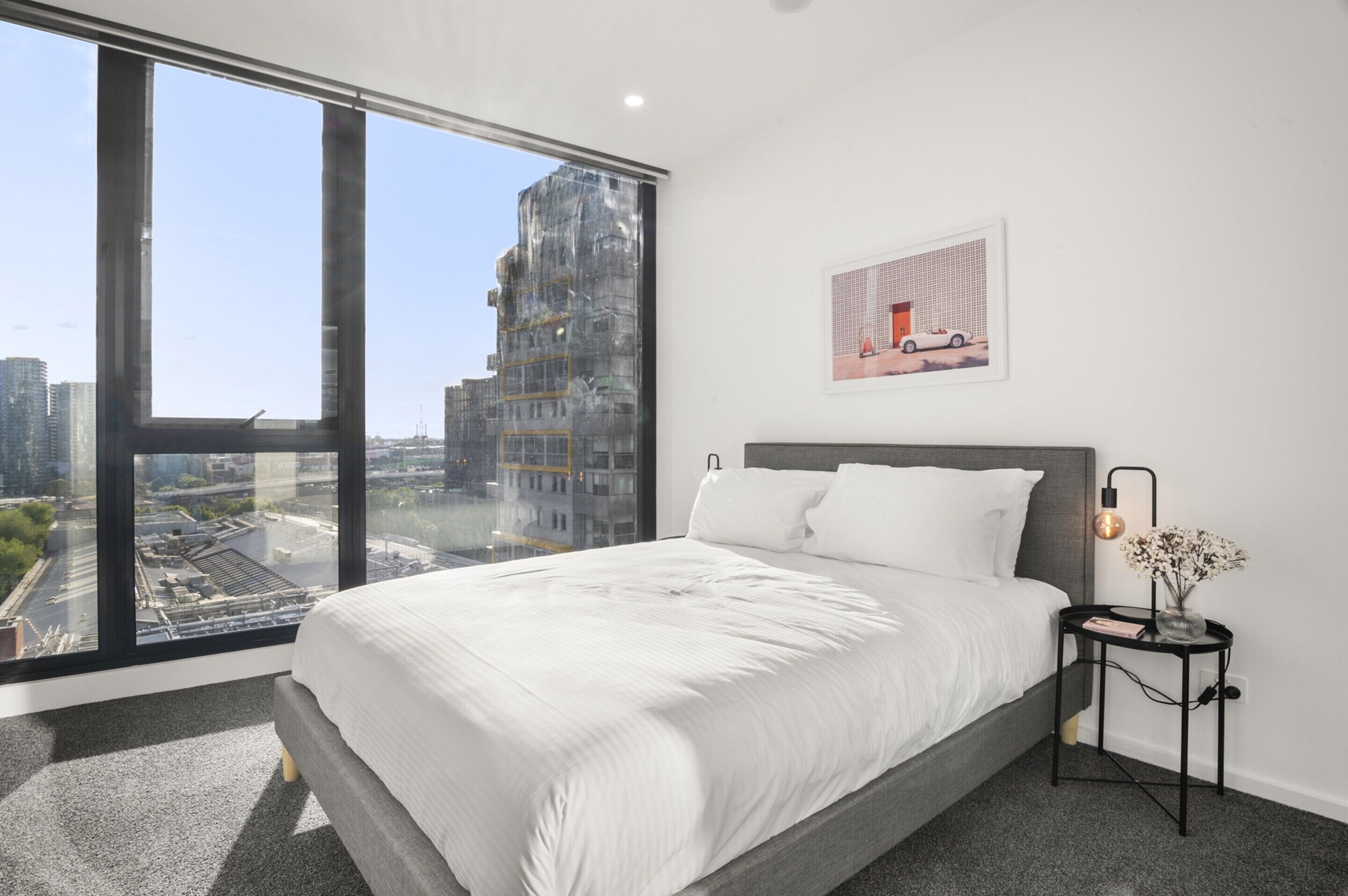 A modern bedroom with a large window, a bed, a nightstand, and a wall-mounted lamp.