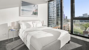A modern hotel room with a large bed, a nightstand, a lamp, and a city view through large windows. - Elegant 2-bed With Superb Views & Amenities (Melbourne)