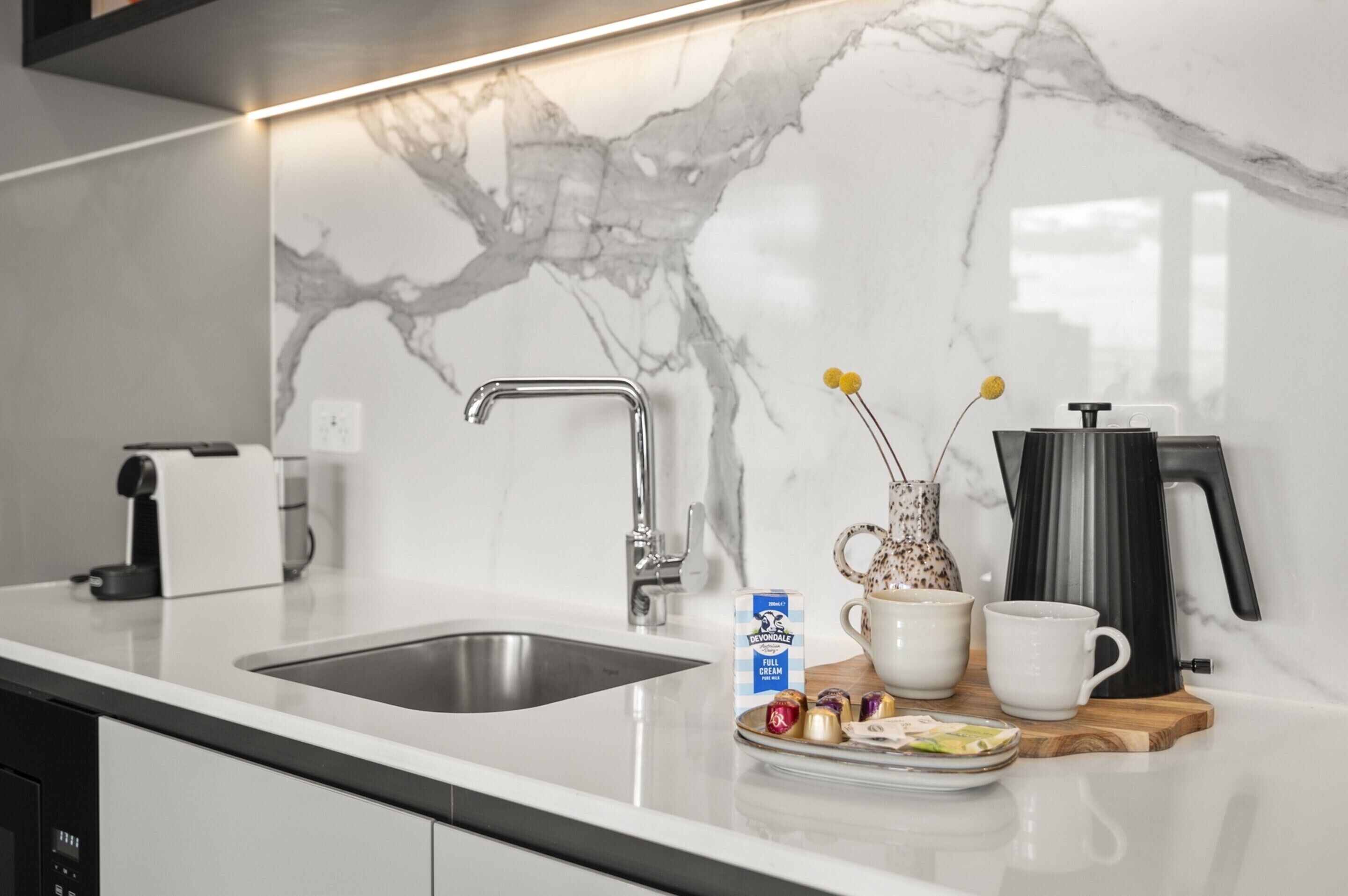 A modern kitchen with a marble backsplash, a stainless steel sink, a coffee maker, a box of Devondale ice cream, and a wooden tray with a cup and a small vase.