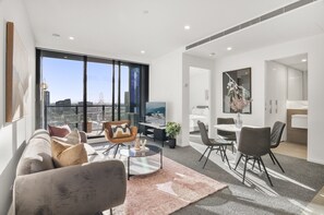 A modern living room with a sofa, coffee table, and chairs, a dining area with a table and chairs, and a large window offering a city view. - Elegant 2-bed With Superb Views & Amenities (Melbourne)