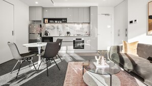 A modern kitchen with grey cabinets, a dining area with a round table and chairs, and a grey sofa with a beige pillow. - Elegant 2-bed With Superb Views & Amenities (Melbourne)