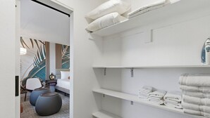 A modern bathroom with white shelves holding towels and a white bathtub. There is a chair and ottoman in the adjacent room. - SVS: Designer Tropical 1BD | Bellagio Fountain View | Resort Pool | Jockey Club (Las Vegas)