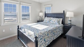 Een slaapkamer met een bed, twee nachtkastjes, een ventilator in het plafond en twee ramen met uitzicht op een strand.