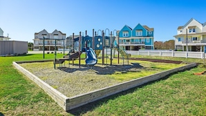 Ein Spielplatz mit Rutsche und Klettergeräten, umzäunt durch einen Holzzaun und mit einer Rasenfläche.