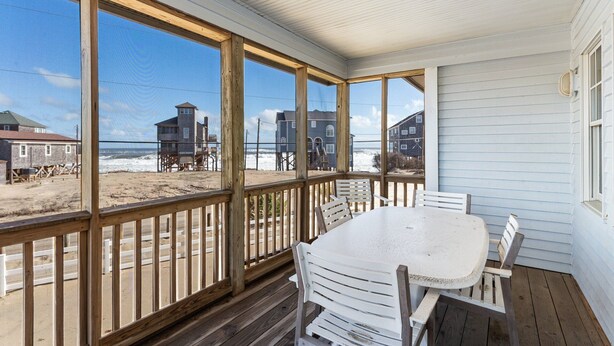 Eine überdachte Veranda mit Tisch und Stühlen, mit Blick auf einen Strand mit Häusern und einem Rettungsturm.